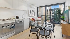Une cuisine moderne dotée d’appareils électroménagers en acier inoxydable, un coin repas avec une table et des chaises en verre, et une grande fenêtre offrant une vue sur la ville.