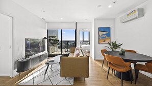 A modern living room with a sofa, a dining table with chairs, a TV, and a large window with a view of the city.