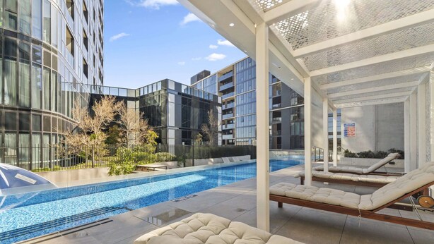 A modern building with a swimming pool, lounge chairs, and a covered seating area.