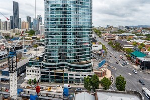 A modern high-rise building with a curved glass facade, surrounded by a busy urban street with construction cranes and vehicles. - Impeccable 2-bed With Pool & Outdoor Dining (Brisbane)