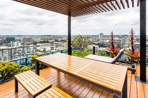 A wooden deck with a lounge chair and bench, overlooking a cityscape with buildings and greenery. - Impeccable 2-bed With Pool & Outdoor Dining (Brisbane)