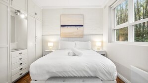 Ein Schlafzimmer mit einem großen Bett, weißen Wänden, einem Gemälde über dem Bett und einem Fenster mit Blick auf Bäume.