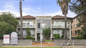 Ein modernes Gebäude mit Betonfassade, großen Fenstern und einem Balkon. Vor dem Gebäude befinden sich Palmen und ein Schild.