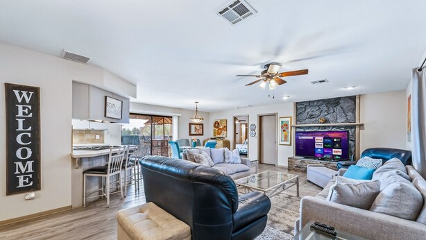 A spacious living room with a black leather sofa, a grey couch, and a glass coffee table. There is a dining area with a white table and chairs, and a kitchen in the background. - HavaHappyStay | Near Lake and Golf~RV/Boat Parking (Lake Havasu City)