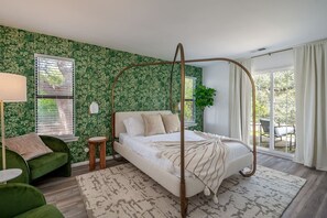 Une chambre à coucher avec un lit à baldaquin, un fauteuil recouvert d’un tissu vert, une petite table d’appoint en bois et une grande fenêtre donnant sur l’extérieur.