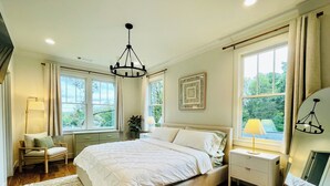 A bedroom with a large bed, a bedside table, a chair, a floor lamp, and a window with curtains. - Grant Park Retreat - Steps to Zoo, & Great Eats (Atlanta)