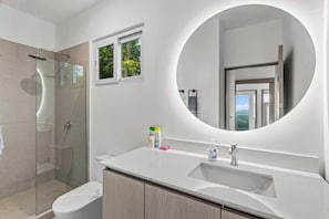 Une salle de bain moderne avec une douche à parois de verre, des toilettes, un lavabo avec un miroir et une fenêtre donnant sur les arbres.