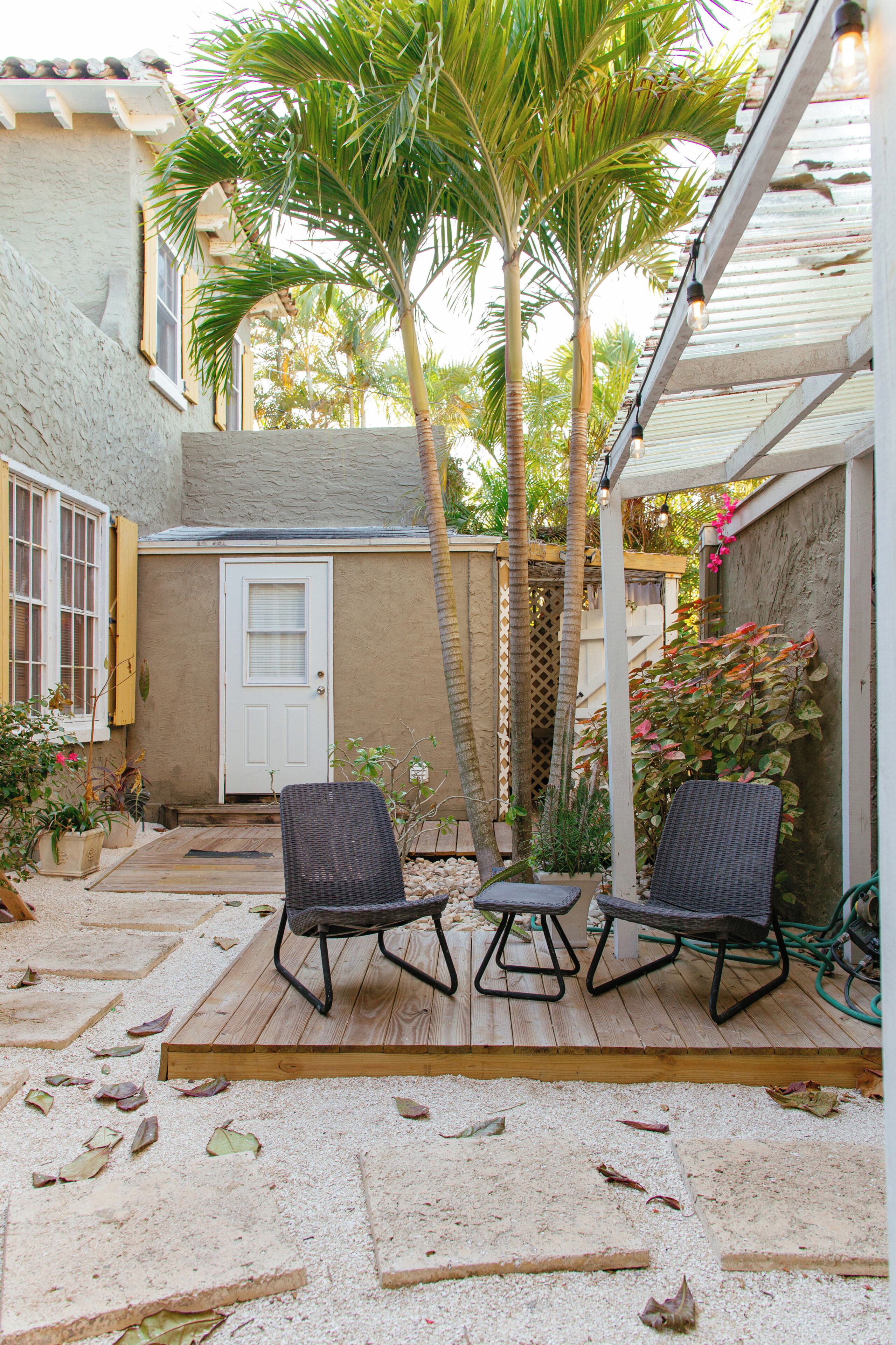 A patio with two wicker chairs, a wooden deck, a door, and a palm tree.