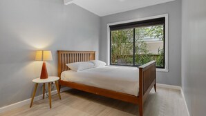 A bedroom with a wooden bed, a bedside table with a lamp, and a large window with a view of greenery outside. - Coastal Retreat on Hazel - Lakes Entrance Getaway (Kalimna)