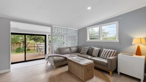 A modern living room with a grey sofa, a coffee table, and a lamp. There is a sliding glass door leading to a patio with outdoor furniture. - Coastal Retreat on Hazel - Lakes Entrance Getaway (Kalimna)