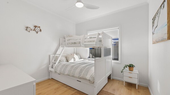 A white room with a bunk bed, a small bedside table, a window, and a ceiling fan.