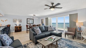 A spacious living room with a large sofa, a coffee table, and a dining area with chairs and a table. A ceiling fan is visible, and there is a view of the ocean through large windows. - Updated 2 bed 2 bath Beachfront Condo with Pool and Amazing Water Views (Indian Shores)