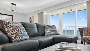 A modern living room with a leather sofa, patterned pillows, a wooden coffee table, and a view of the ocean through large windows. - Updated 2 bed 2 bath Beachfront Condo with Pool and Amazing Water Views (Indian Shores)