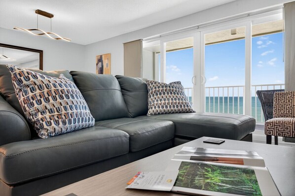 A modern living room with a leather sofa, patterned pillows, a wooden coffee table, and a view of the ocean through large windows.