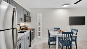 A modern kitchen with stainless steel appliances, a dining area with blue chairs, and a flat-screen TV mounted on the wall. - Your Gulf Coast Escape in Madeira Beach (St. Petersburg)