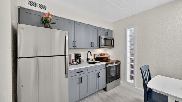 A modern kitchen with grey cabinets, a stainless steel refrigerator, a microwave, a coffee maker, and an oven. There is a dining table with chairs and a window with blinds. - Your Gulf Coast Escape in Madeira Beach (St. Petersburg)