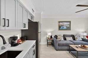 A modern kitchen with white cabinets, a built-in oven, and a coffee maker. There is a gray sofa with patterned cushions in the living area, and a painting on the wall. - Ground Floor Stunner Steps From the Beach (St. Petersburg)
