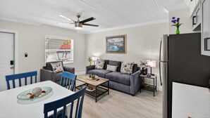 A modern living room with a grey sofa, a dining table with blue chairs, a ceiling fan, and a refrigerator. - Coastal Comfort Just Steps from the Sand (St. Petersburg)