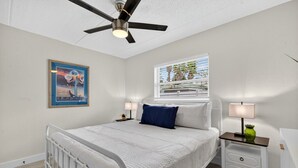 Ein Schlafzimmer mit Bett, Deckenventilator, Fenster mit Aussicht, einem gerahmten Bild an der Wand und einem Nachttisch mit Lampe.