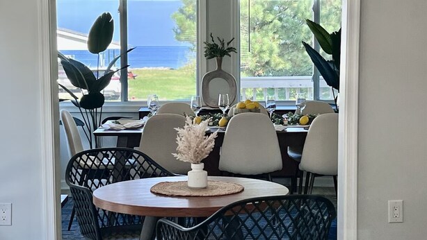 An indoor dining area with a view of the ocean, featuring a round table with chairs, a vase with dried flowers, and a potted plant.