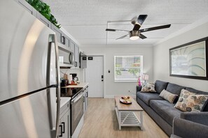 Ett modernt kök med ett inbyggt kylskåp, en mikrovågsugn och en kaffebryggare på bänkskivan. I vardagsrummet finns en grå soffa med mönstrade kuddar, och en takfläkt hänger ovanför.