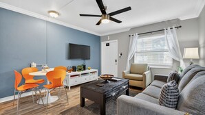 Un salon doté d’un ventilateur de plafond, d’un téléviseur fixé au mur, d’un coin repas avec des chaises orange, d’un canapé et d’une fenêtre avec des rideaux.
