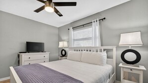 Une chambre à coucher comprenant un lit, un téléviseur, un ventilateur de plafond, une fenêtre avec des rideaux et une table de chevet avec une lampe.
