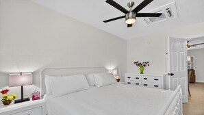 A white bedroom with a bed, bedside tables, a nightstand, a green vase with flowers, a ceiling fan, and a door leading to another room.