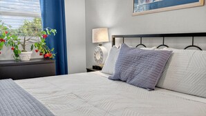 Una cama bien hecha con un colcha estampada, una lámpara de noche y una pequeña maceta con flores rojas sobre una mesita.