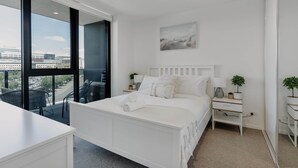 A modern bedroom with a large bed, a bedside table, a nightstand, and a view of the city through large windows. - Cosy 1-bed With Pool, Gym ,parking (Canberra)