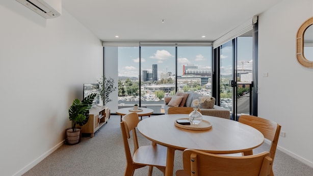 A modern living room with a round wooden table, comfortable seating, a large window with a city view, and a potted plant. - Cosy 1-bed With Pool, Gym ,parking (Canberra)