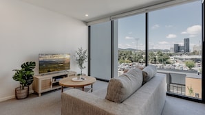 A modern living room with a large window, a flat-screen TV, a sofa, a coffee table, and a potted plant. - Cosy 1-bed With Pool, Gym ,parking (Canberra)