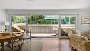 A spacious living room with a wooden dining table, chairs, and a woven sofa. Large windows offer a view of the outdoors. - Poolside Stay - Close to Taronga Zoo & Luna Park (Sydney)