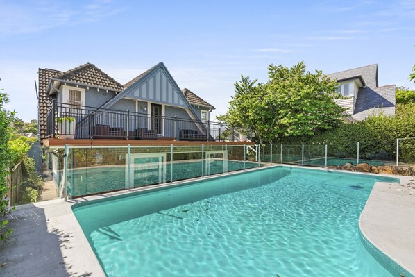 A swimming pool with a clear blue water surface, surrounded by a fence and a house with a sloped roof.