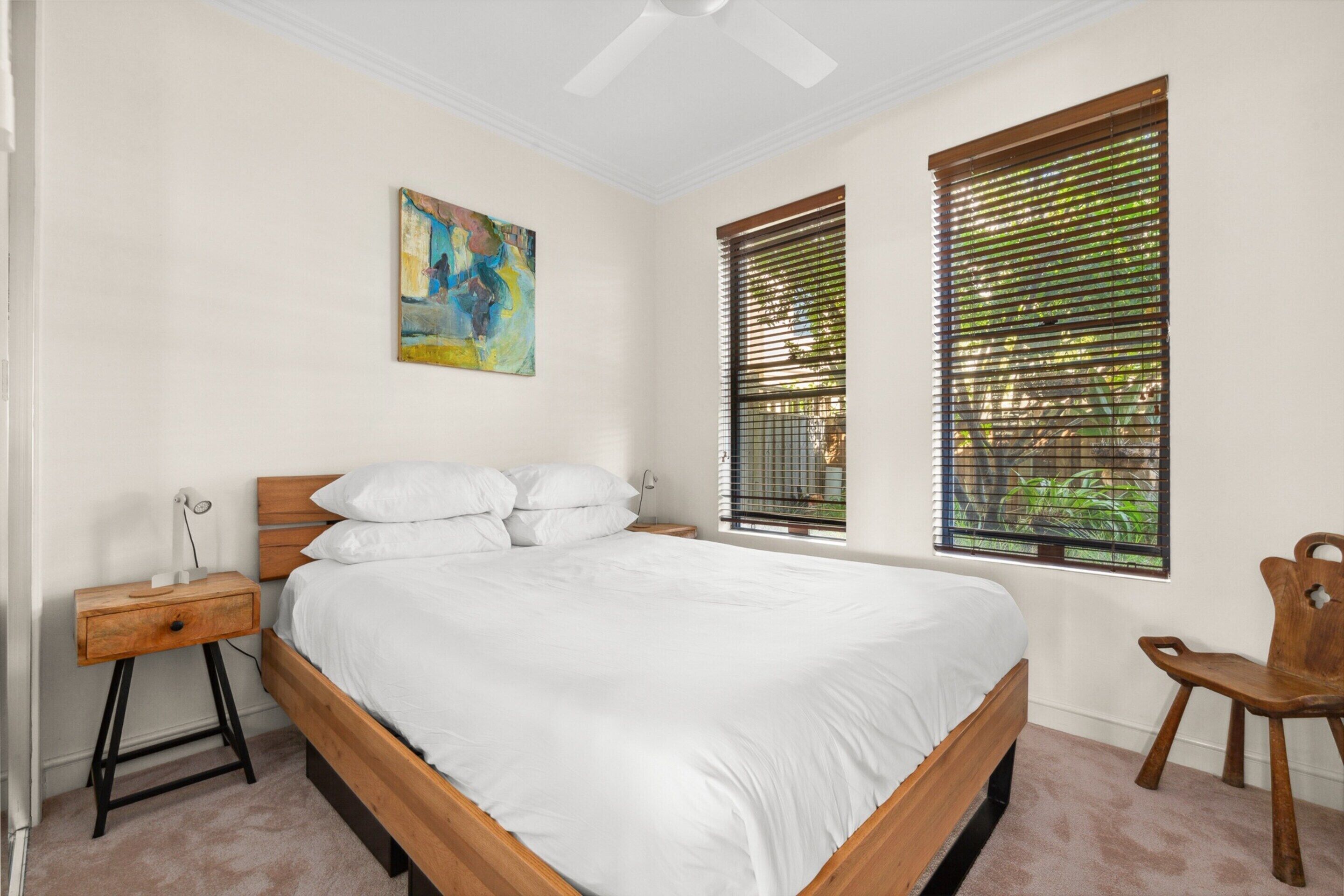 A bedroom with a wooden bed, white bedding, a nightstand, a chair, and a painting on the wall.