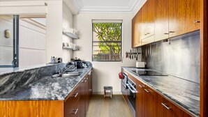 A modern kitchen with wooden cabinets, stainless steel appliances, a marble countertop, and a window with a view of greenery. - Elegant Stay in Balmain With Pool Access (Sydney)