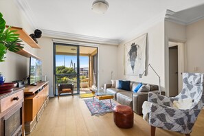 A modern living room with a sofa, armchair, and coffee table. There is a kitchen area with a dining table and chairs. A large window provides a view of the outdoors. - Elegant Stay in Balmain With Pool Access (Sydney)