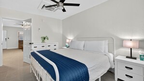 Un dormitorio con cama, mesitas de noche, ventilador de techo y puerta que da a otra habitación.