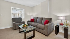 Una sala de estar con un sofá gris, un sillón gris, una mesa de centro de vidrio y una mesita auxiliar de vidrio con una lámpara.