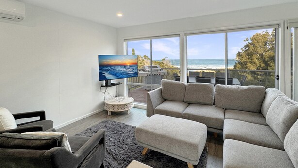 A modern living room with a large TV, grey sofa, and a view of the ocean through large windows.