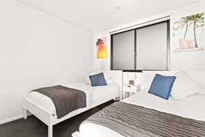 A hotel room with two single beds, a large window, a flat-screen TV, and a small bedside table with a lamp. - Ocean Views, Deck and Parking at Beach Apartment (Sydney)