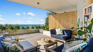 A spacious outdoor seating area with a wooden deck, a sofa, and a bookshelf. - Ocean Views, Deck and Parking at Beach Apartment (Sydney)