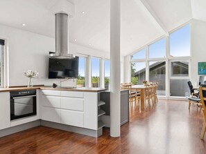A modern kitchen with a built-in oven and a large TV. There is a dining area with wooden chairs and a table, and large windows offering a view of the outdoors. - 4 Star Holiday Home in Holbaek (Holbaek)