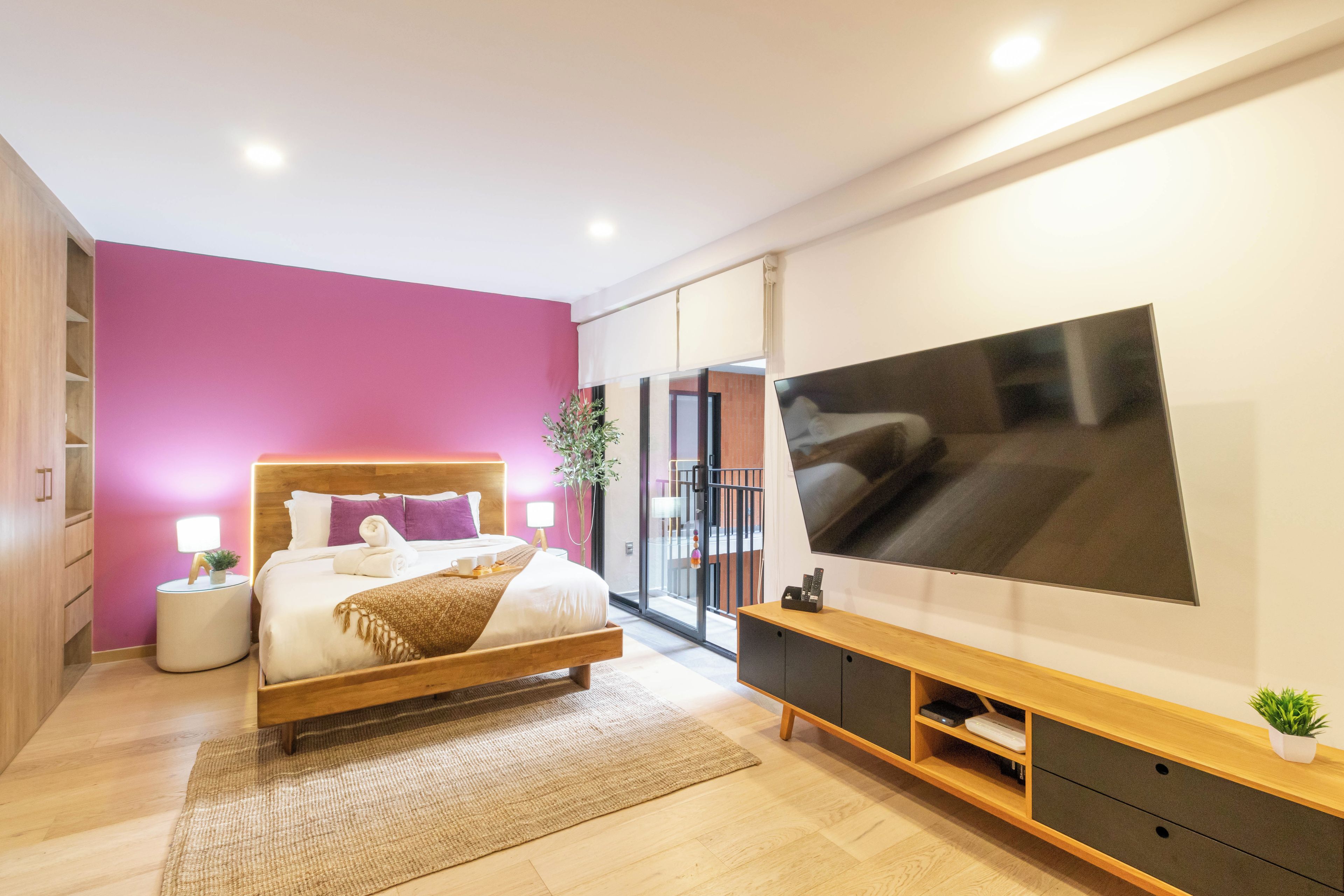 A modern bedroom with a large bed, a flat-screen TV, a bedside table with a lamp, and a small plant.