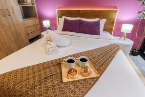 Una habitación de hotel con una cama, una mesita de noche, una lámpara y una bandeja con dos tazas de café y un pastel. También hay una toalla sobre la cama.