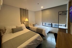 A hotel room with two beds, a bedside table, a lamp, a TV, and a window with a view. - AKELA 30 HOTEL (Medellín)