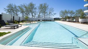 Uma piscina com espreguiçadeiras e vista para árvores e um edifício ao fundo.
