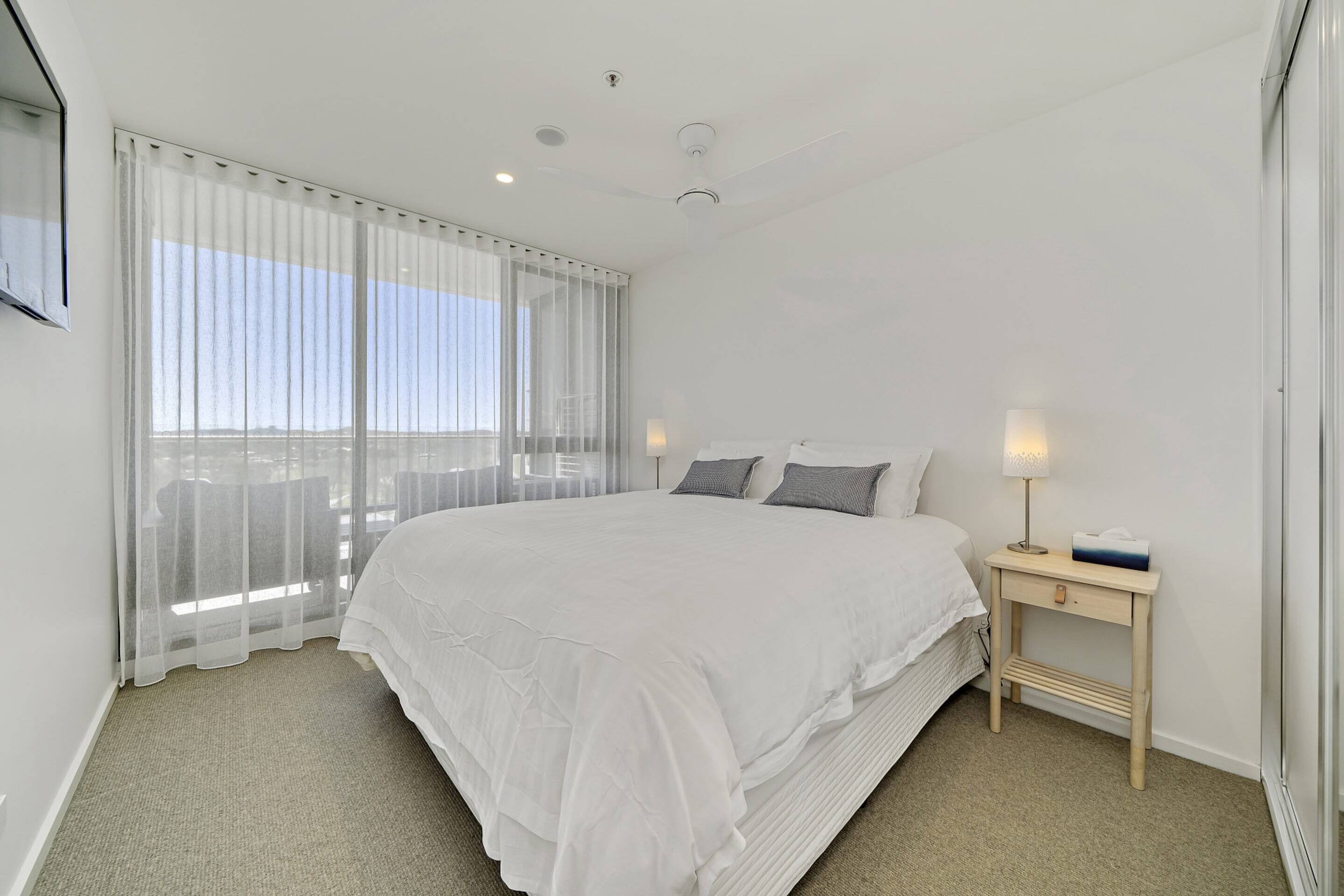 A hotel room with a large bed, a bedside table with a lamp, and a view of the city through sheer curtains.