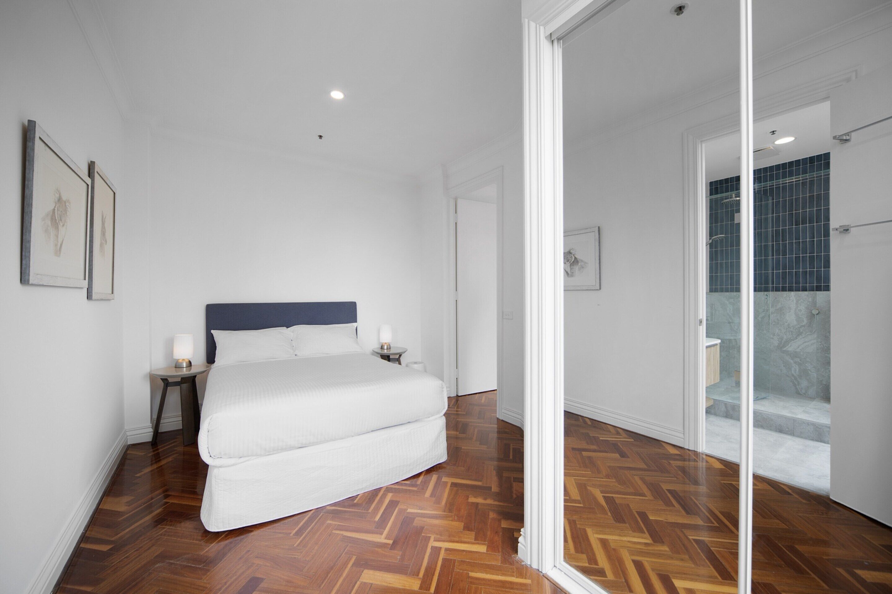 A bedroom with a large bed, wooden flooring, a nightstand, and a wall-mounted lamp.