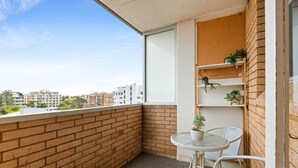 Ein Balkon mit Ziegelwand, Glasdoor und einem kleinen Tisch mit Stühlen. Auf einem Regal stehen Blumentöpfe.
